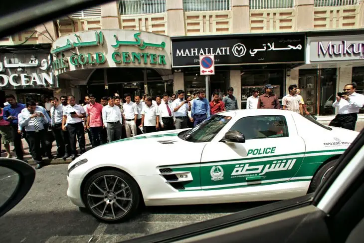 Patrol passing by the street and people on the side of a street watching sports cars 